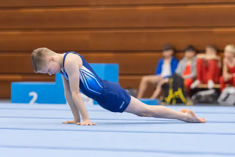 Young gymnast in blue leotard holds a plank position on floor exercise mat, teammates watching from bleachers behind