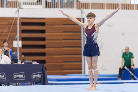 Young gymnast in maroon and navy leotard stands with arms raised in finishing pose on blue competition mat