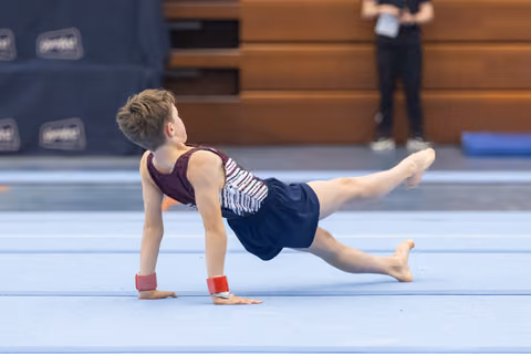 Young gymnast performs a plank-based floor exercise position with one leg extended, wearing navy and white leotard on blue mat