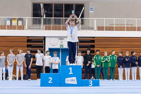 Young gymnast raises trophy overhead on first place podium while teammates stand below in gymnasium