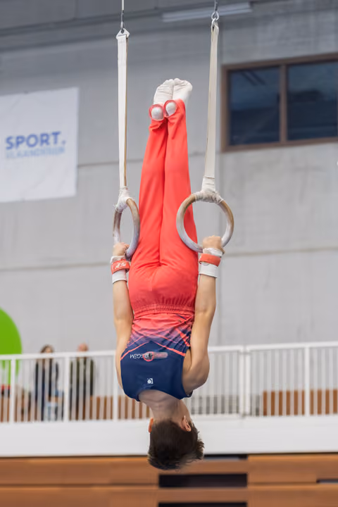 Young gymnast performing an inverted hang on rings, body fully extended upside down in red leotard at indoor facility