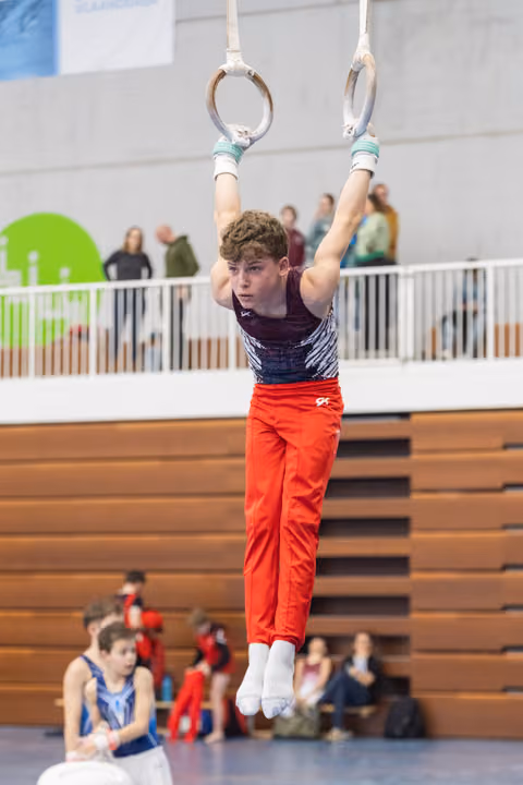 Young gymnast in red pants hanging from still rings with focused expression, spectators watching from bleachers