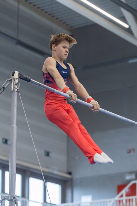 Young gymnast performing on horizontal bar with intense concentration, wearing red pants and navy leotard in training facility