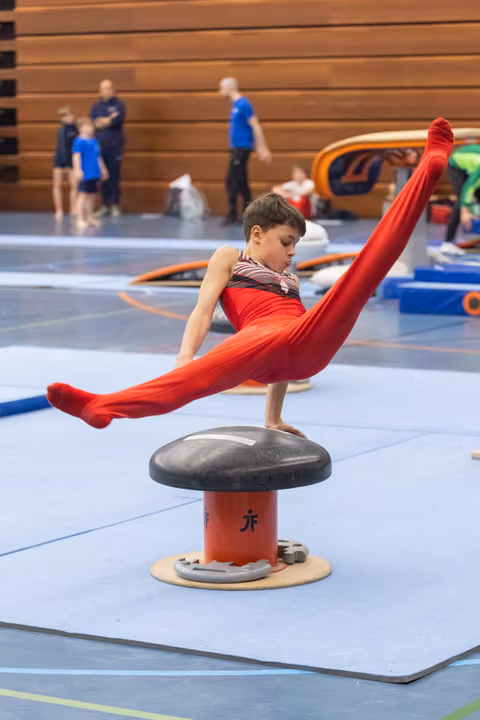 Young gymnast in red performs extended scissor position on pommel horse during training in indoor gymnasium