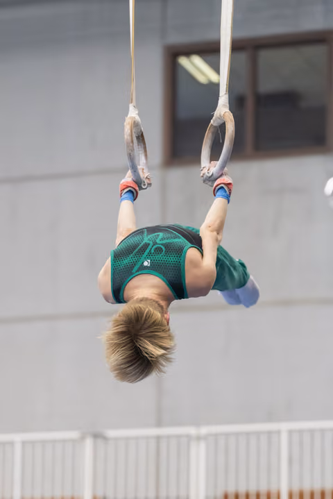 Young gymnast performing inverted hang on still rings, body arched backward in green leotard during apparatus routine