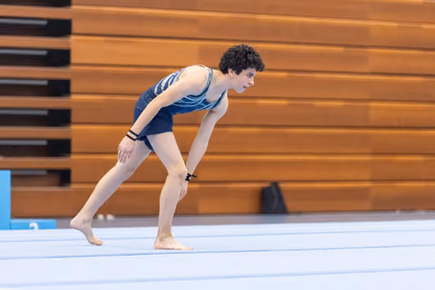 Young male gymnast in blue leotard performs a deep lunge on floor mat, demonstrating flexibility and concentration