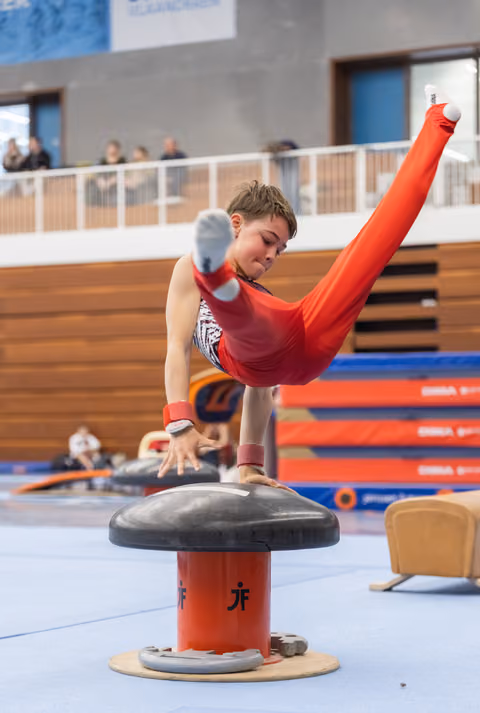 Male gymnast in red leotard executes a horizontal scissor move on pommel horse, demonstrating strength and control