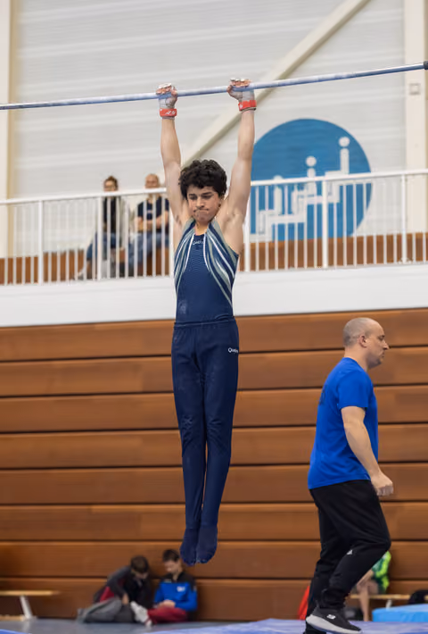 Young gymnast in navy uniform hangs from high bar with coach observing, demonstrating strength in training facility