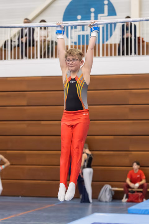 Young gymnast in red pants and colorful leotard hanging from horizontal bar, arms raised, determined expression behind glasses