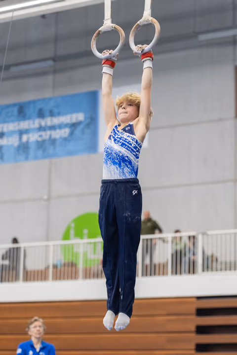 Young gymnast hanging from rings with focused expression, wearing blue and white patterned leotard in indoor training facility