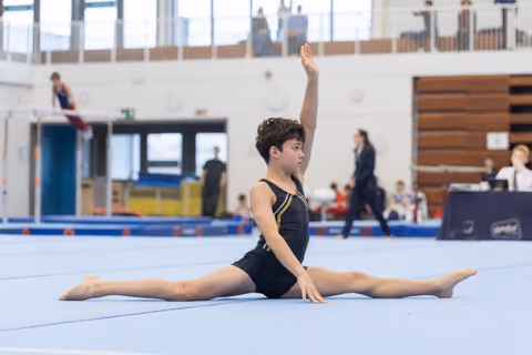 Young gymnast in black leotard performs a full split with one arm raised, demonstrating flexibility on the blue competition floor