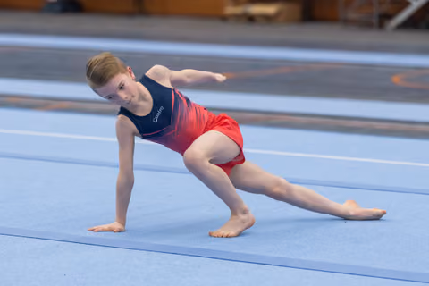 Young gymnast performs a side lunge pose during floor routine, displaying flexibility and pointed toes on blue mat