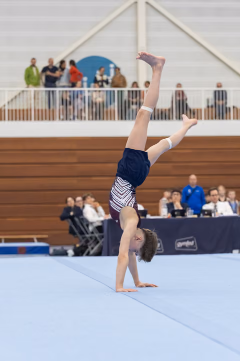 Young gymnast executes a handstand during floor routine, legs extended vertically, in front of judges and spectators
