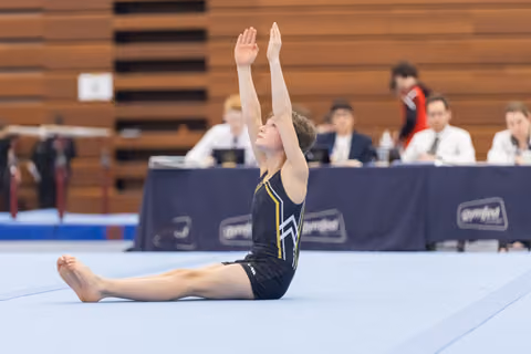 Young gymnast in black leotard performs seated stretch with arms raised during floor routine at gymnastics meet