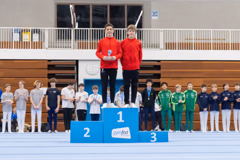 Two young gymnasts in red tracksuits stand on the first place podium holding a trophy, with other competitors on lower podiums