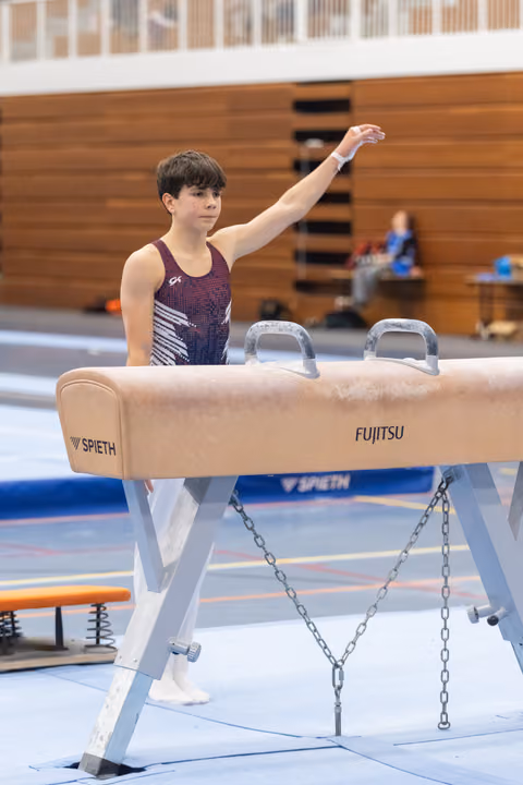Young gymnast raises arm in salute beside Fujitsu pommel horse during training session in wooden gymnasium