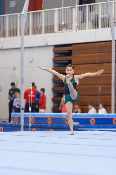 Young gymnast performs arabesque pose on floor exercise mat, arms extended, displaying balance and grace in teal leotard