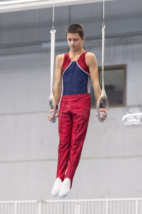 Young gymnast performing on still rings, focused expression, wearing red and navy uniform in training facility