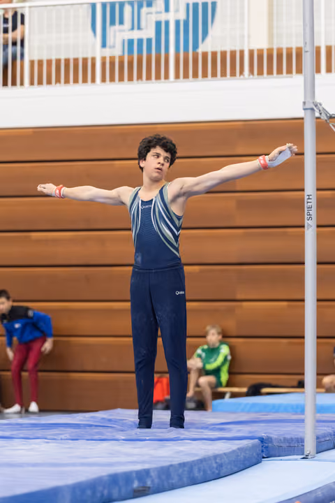 Young gymnast in navy leotard stands with arms outstretched in preparation pose on mat, focused expression before routine