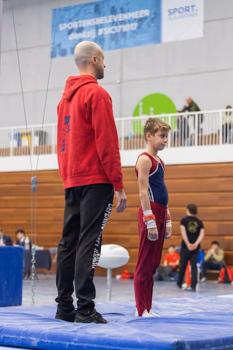Coach in red hoodie stands beside young gymnast in maroon uniform on blue floor mat, observing the arena together