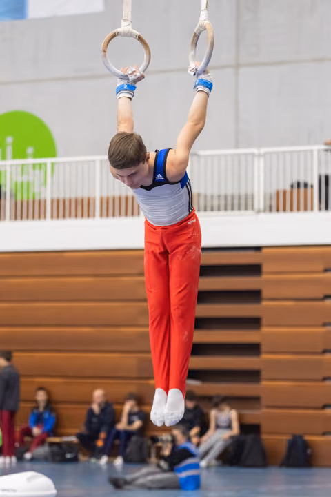 Young gymnast in red pants and white-blue leotard performing a hang position on still rings during indoor meet
