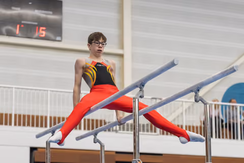 Young gymnast executes a split position on parallel bars, wearing red pants and black-orange top, focused expression