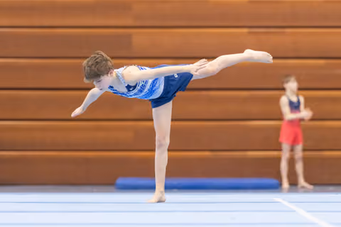 Young gymnast performs an arabesque balance on floor exercise while another athlete waits in the background