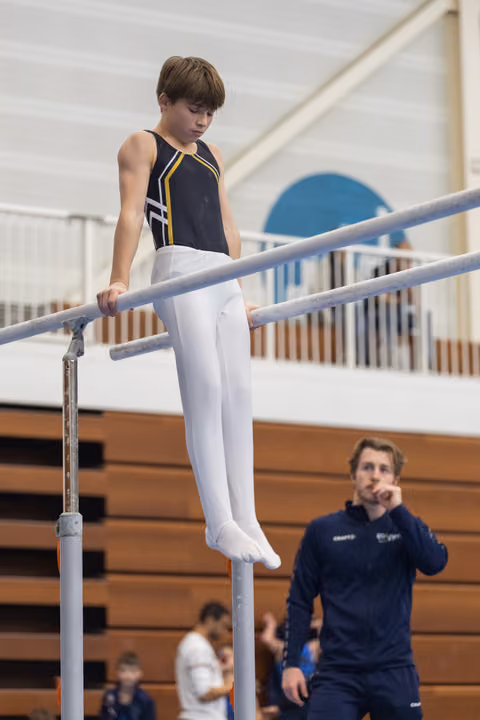 Young gymnast in navy leotard performs on parallel bars with focused concentration while coach observes from bleachers