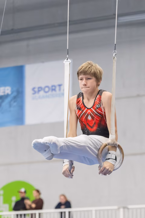 Young gymnast performing L-sit position on rings, displaying focused concentration in red and black leotard at indoor facility