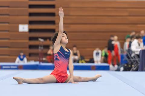 Young gymnast in pink-navy leotard performs floor split with arm raised elegantly, displaying flexibility in competition venue
