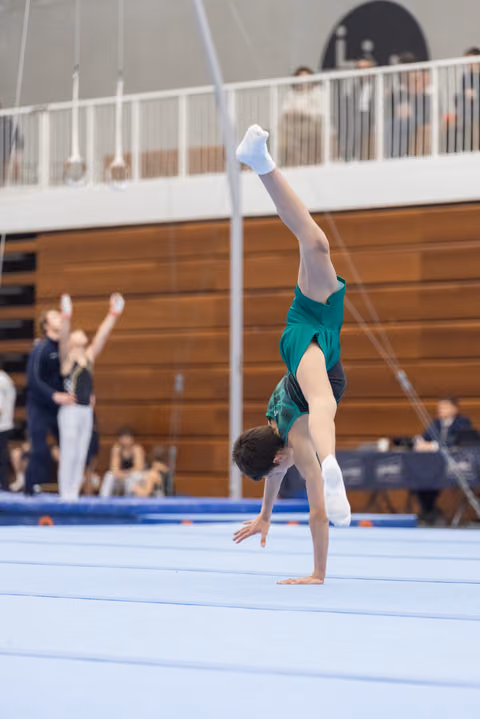 Gymnast performing handstand on floor exercise mat in green leotard, legs extended upward, spectators visible in background