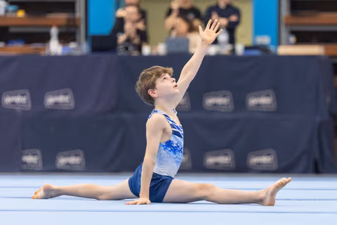 Young gymnast performs full splits on floor with dramatic arm extension, showcasing flexibility and artistic expression