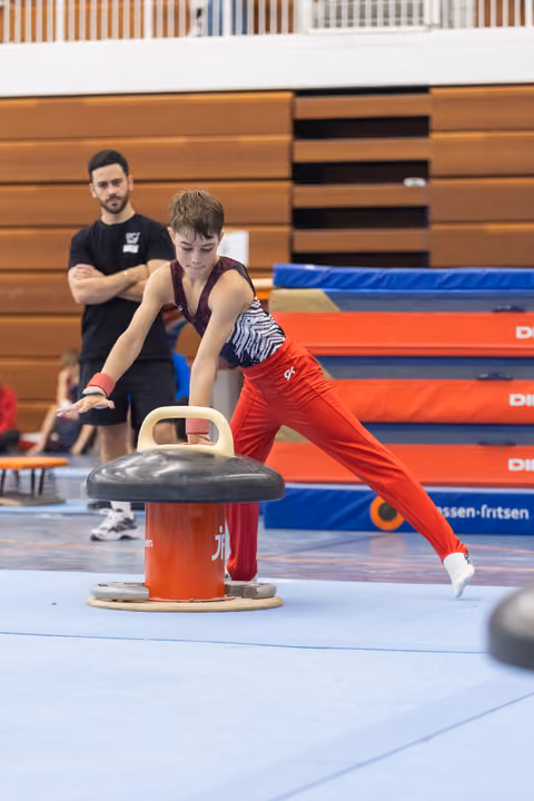 Young gymnast in red pants practices pommel horse technique while coach observes in background at training facility