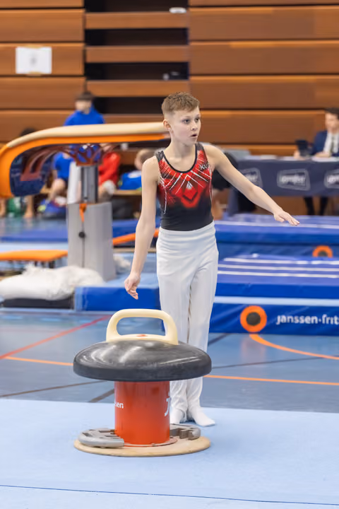 Young male gymnast in red and black leotard stands beside mushroom apparatus, arms extended, focused expression during training