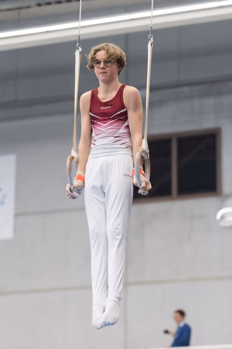 Gymnast suspended on still rings with focused expression, wearing gradient burgundy leotard and white pants in training facility