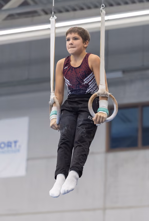 Young male gymnast holds position on rings apparatus, wearing burgundy and black leotard with focused expression
