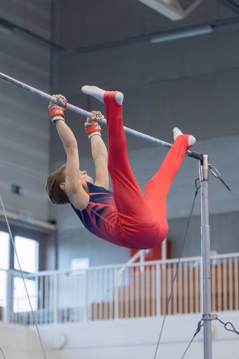 Young gymnast in red outfit performs inverted hang on high bar, legs extended upward in indoor training facility