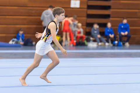 Young gymnast in white leotard running barefoot across blue floor mat during routine, spectators visible in background
