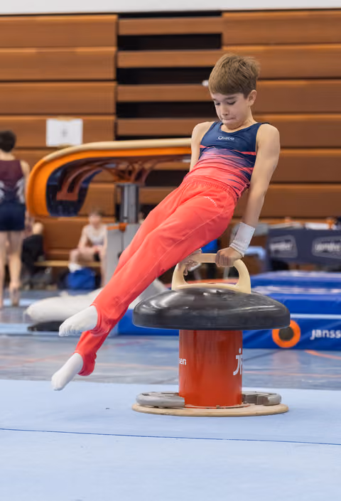 Young gymnast in red pants and blue leotard performing a strength hold on the pommel horse mushroom apparatus