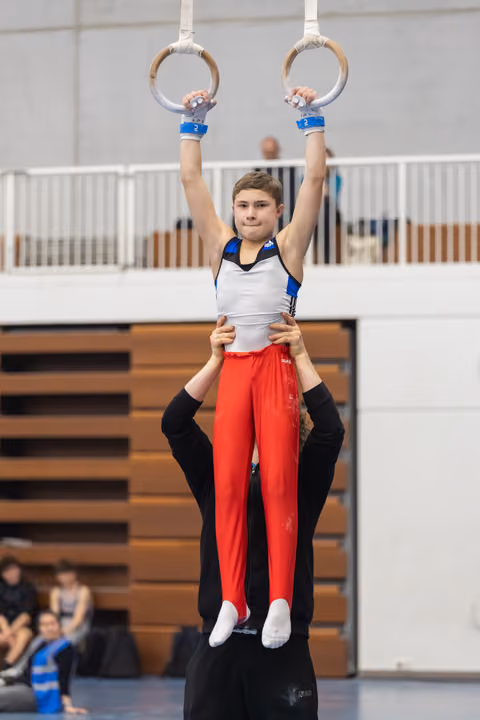 Young gymnast in red pants hangs from rings while coach supports from below in gymnasium