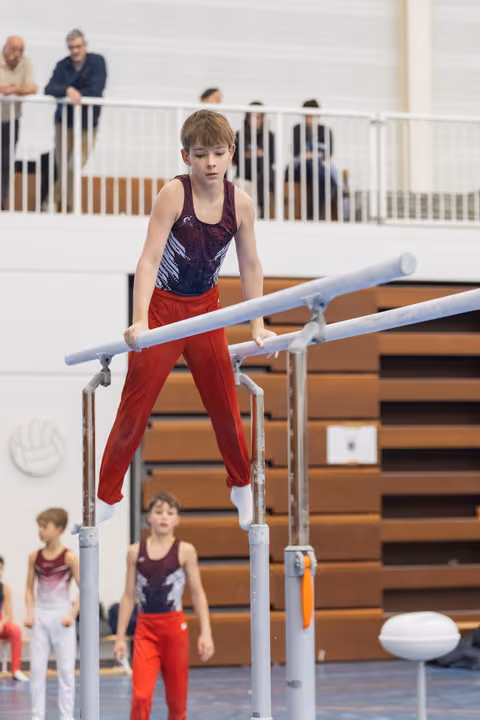 Young gymnast in red pants and purple leotard performs on parallel bars with focused concentration while teammates watch below