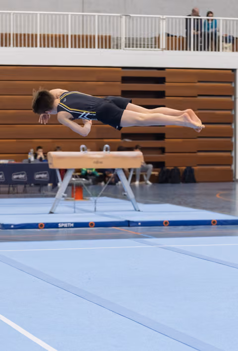 Gymnast performs horizontal backflip above vault, body fully extended in mid-air during routine in gymnasium