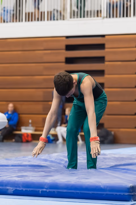 Male gymnast in green unitard bends forward on blue mat during floor routine, showing focused concentration in gym setting