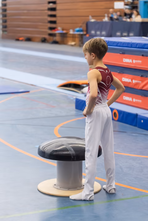 Young gymnast in burgundy leotard and white pants stands with hand on hip beside a mushroom trainer in the gym