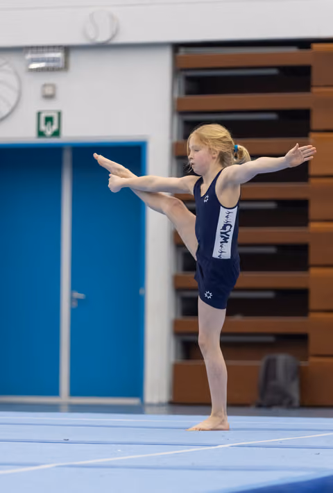 Young gymnast in navy leotard performs a standing split with arms extended, demonstrating flexibility and balance on floor mat
