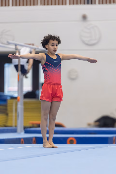 Young gymnast with curly hair performing a T-pose balance on floor mat, arms extended, displaying focused concentration