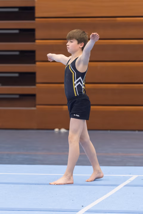 Young gymnast performs floor routine with arms extended, demonstrating focused form on blue mat in gymnasium