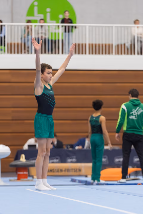Gymnast in green and navy leotard stands with arms raised overhead, completing a floor routine at an indoor gymnastics meet.