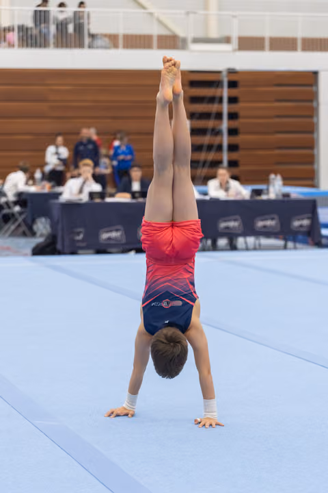 Young gymnast performs a handstand on blue floor mat, legs straight up, with judges' table and wooden wall in background