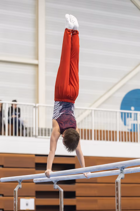 Young gymnast performs a vertical handstand on parallel bars, wearing red top and patterned shorts in indoor training facility